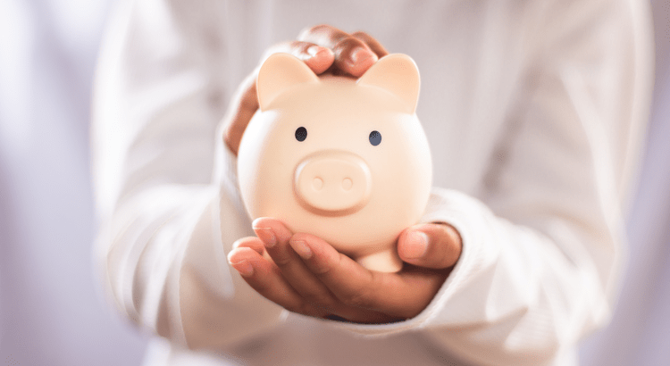 Person holding a small beige piggy-bank in both hands, symbolizing budgeting and home-selling savings.