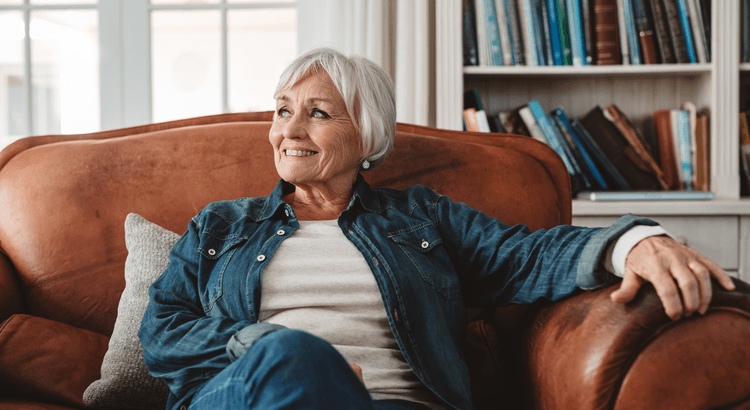 Smiling older woman relaxing in a leather chair inside a cozy living room with bookshelves in the background.